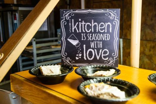 A sign reads "This kitchen is seasoned with love" next to plates of food on a wooden counter.