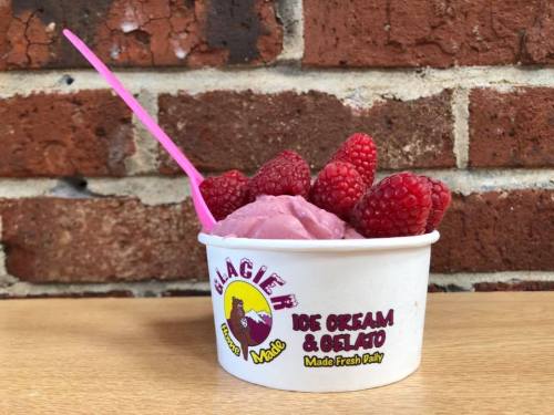 A cup of raspberry ice cream topped with fresh raspberries, set against a brick wall background.