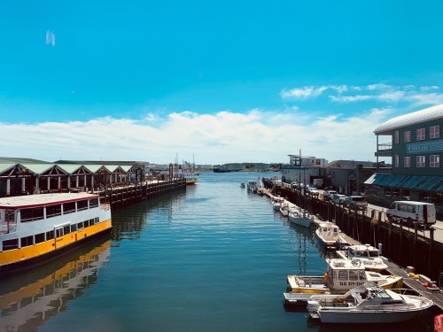 A sunny harbor scene with boats docked along the pier and buildings lining the waterfront under a clear blue sky.
