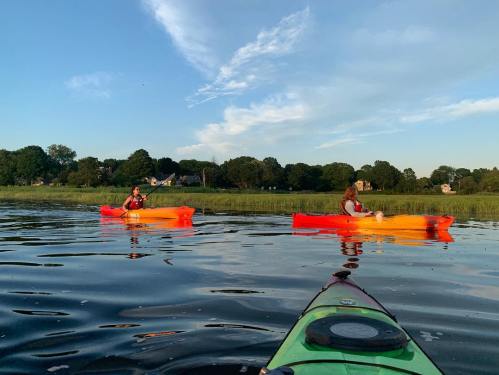 Two people kayak on calm water, surrounded by greenery and a clear blue sky, with one kayak in the foreground.
