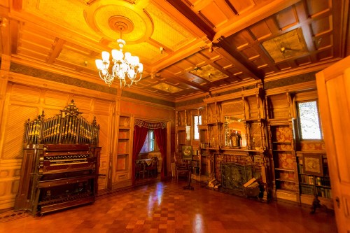 A warmly lit vintage room featuring an ornate organ, bookshelves, and a decorative fireplace.