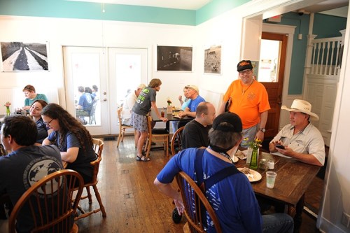 A busy café with people dining, chatting, and a woman serving, featuring artwork on the walls and a bright atmosphere.