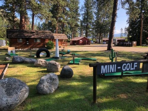A mini-golf course with green obstacles and rocks, surrounded by trees and cabins in a scenic outdoor setting.