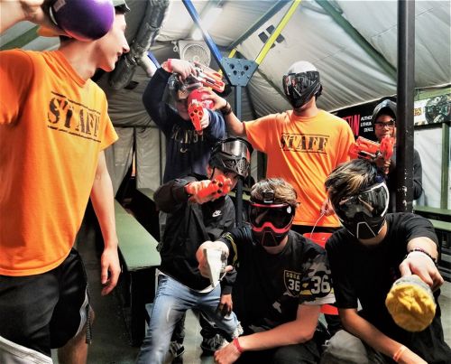 A group of people in masks and protective gear pose with toy guns in a paintball arena. Staff members are highlighted.