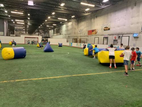 Children playing in an indoor arena with inflatable obstacles, engaged in a game of paintball or similar activity.