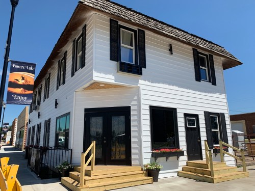 A two-story white building with black shutters, wooden steps, and a sign reading "Powers Lake Enjoy" on a sunny day.