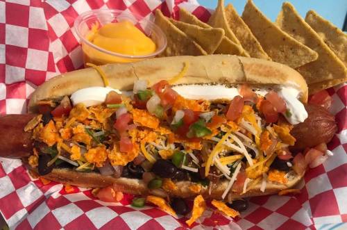 A loaded hot dog topped with cheese, tomatoes, black beans, and crushed chips, served with tortilla chips and cheese dip.