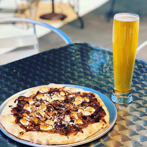A pizza topped with mushrooms and almonds on a metal table, next to a tall glass of golden beer.
