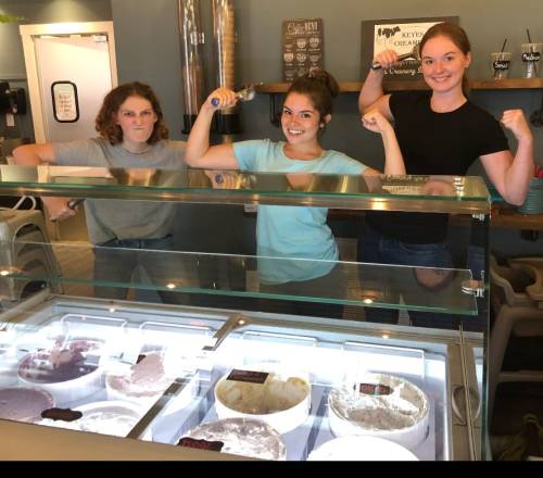 Three women flex their muscles playfully in front of an ice cream display, showcasing their strength and fun spirit.