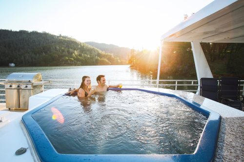 A couple relaxes in a hot tub on a boat, surrounded by scenic mountains and a sunset over the water.