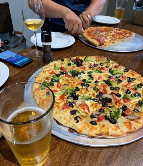 A table with two pizzas, a glass of beer, and a wine glass, with a person preparing to serve pizza.