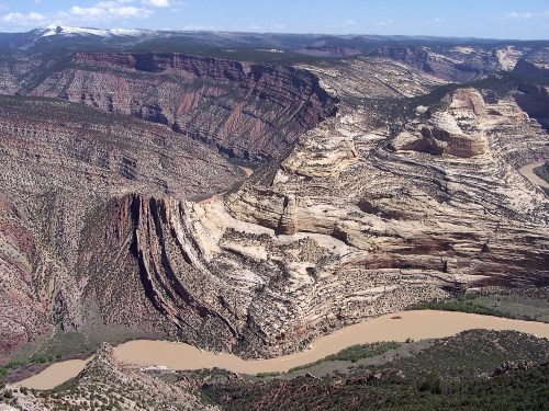 Aerial view of rugged, layered rock formations and a winding river in a vast canyon landscape under a clear blue sky.