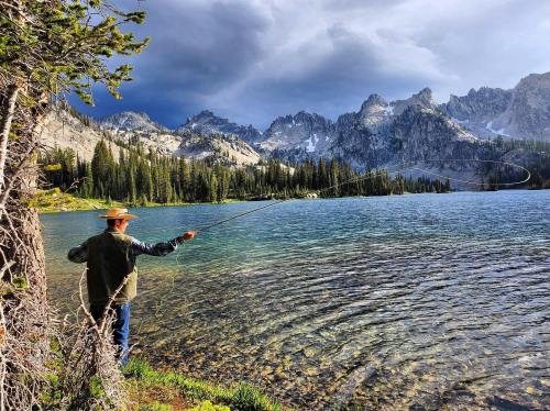A person fishing by a serene lake, surrounded by mountains and lush trees under a partly cloudy sky.