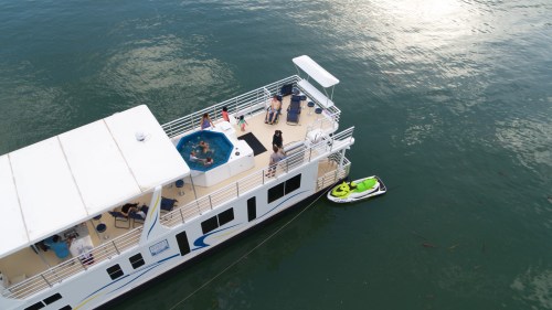 Aerial view of a houseboat with people enjoying a hot tub and a jet ski nearby on calm water.