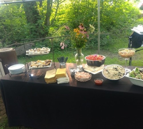 A buffet table outdoors featuring various dishes, flowers, and utensils, set against a green backdrop.