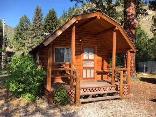 A cozy wooden cabin with a porch, surrounded by trees and gravel, set against a mountainous backdrop.