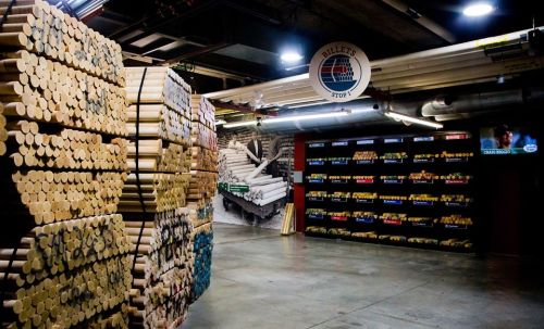 A warehouse interior filled with stacked wooden billets and a wall of colorful materials, with a TV in the background.
