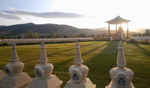 A serene landscape featuring stone stupas, lush green grass, and a pagoda against a backdrop of mountains at sunset.