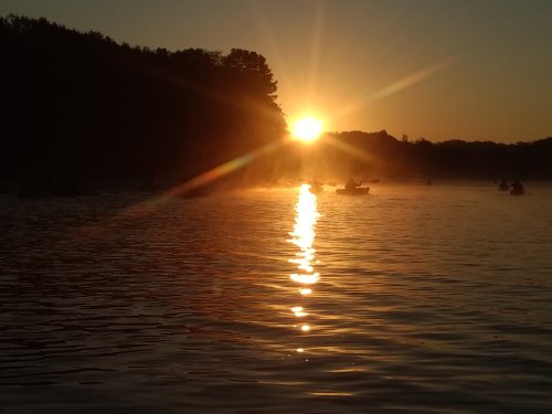 Sunset over a calm lake, with silhouettes of kayakers and mist rising from the water.