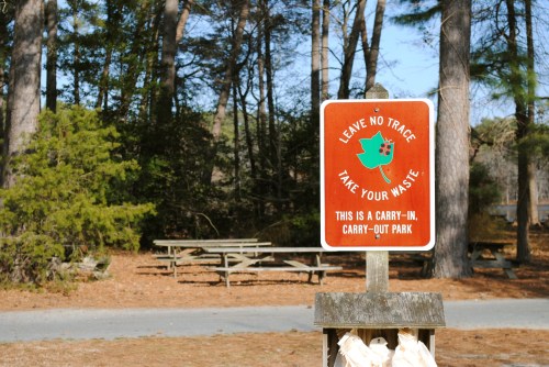 A sign in a park reads "Leave No Trace, Take Your Waste" with a green leaf graphic, surrounded by trees and picnic tables.