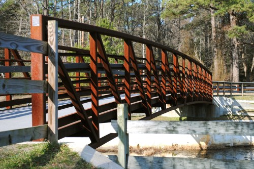 A curved metal bridge with wooden railings spans a small waterway, surrounded by trees and greenery.