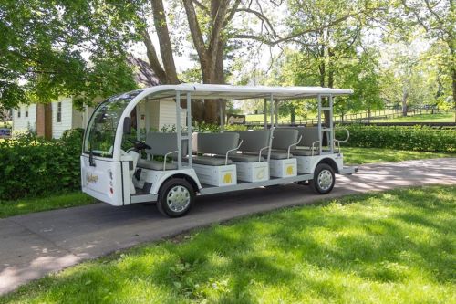 A white electric shuttle with gray seats parked on a grassy path surrounded by trees and greenery.