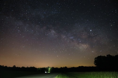 A starry night sky filled with the Milky Way, above a dark road bordered by trees and grass.