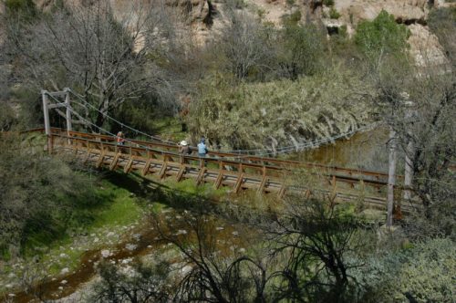 A wooden suspension bridge spans a creek, with two people walking across surrounded by trees and rocky terrain.