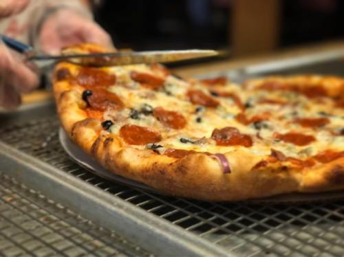 A hand using a knife to slice a pepperoni pizza with olives and cheese on a metal tray.