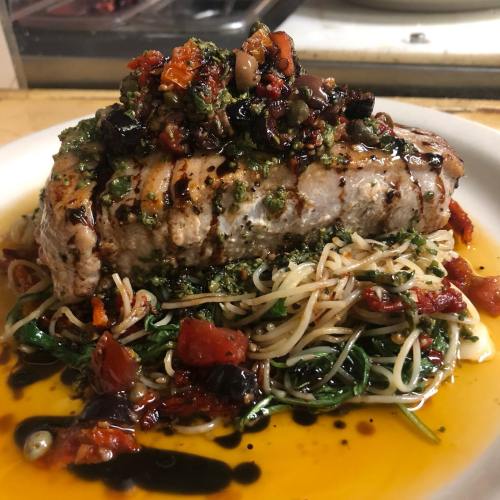 A plate of grilled fish topped with a colorful salsa, served over a bed of pasta and sautéed greens in olive oil.
