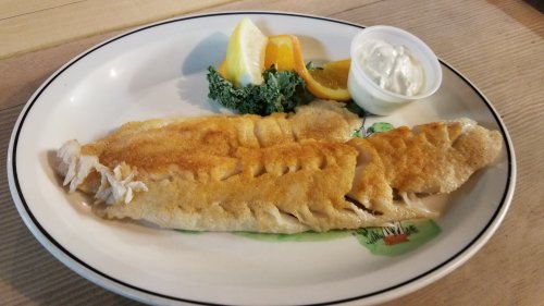 A plate of fried fish fillets with lemon wedges, orange slices, and a side of tartar sauce on a wooden table.