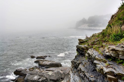 Foggy coastal scene with rocky shoreline and gentle waves, creating a serene and mysterious atmosphere.