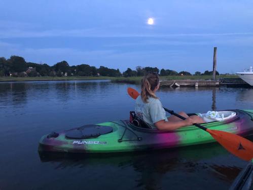 A person in a kayak on calm water at dusk, with a full moon reflecting above and a dock in the background.