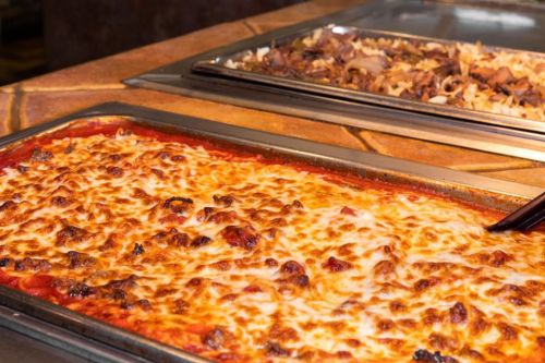 A tray of cheesy baked pasta with marinara sauce, next to a tray of seasoned meat and rice.