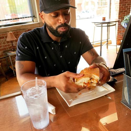 A man sits at a table in a restaurant, holding a large sandwich and looking at it with interest. A glass of water is nearby.