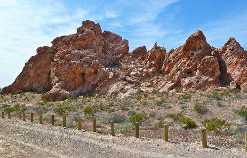 Rocky formations rise against a blue sky, surrounded by sparse vegetation and a dirt road lined with wooden posts.