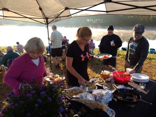 A group of people enjoying a picnic by a misty lake, with one woman serving food from a table under a canopy.