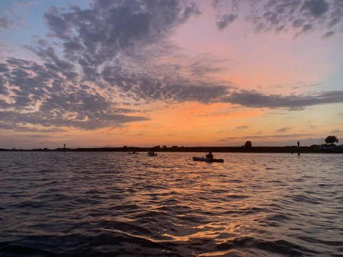 A serene sunset over water, with colorful clouds reflecting on the surface and silhouettes of boats in the distance.