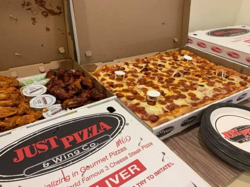 A spread of pizza and chicken wings in takeout boxes from Just Pizza & Wing Co. with plates and dipping sauces.