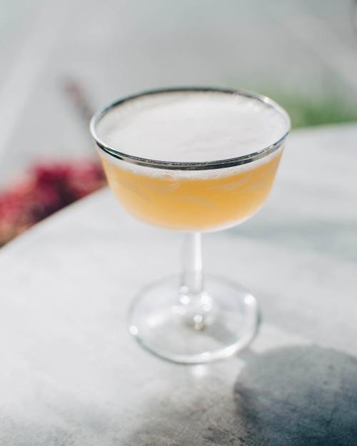 A frothy cocktail in a delicate glass sits on a table, with a blurred outdoor background.
