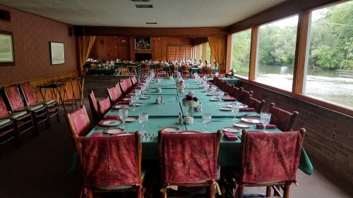 A spacious dining room with long tables set for a meal, overlooking a river through large windows.