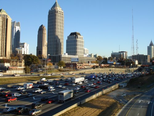 Busy highway with heavy traffic, surrounded by tall buildings and skyscrapers in an urban setting. Clear blue sky above.