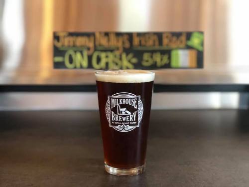 A pint of dark amber beer with a frothy head, featuring a Milkhouse Brewery logo, set against a chalkboard menu.