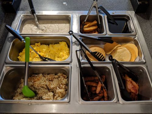 A buffet station with various food items: scrambled eggs, pancakes, bacon, sausage, hash browns, and sauces.