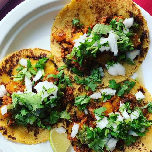 Three tacos on a plate, topped with chopped onions and cilantro, with a lime wedge on the side.