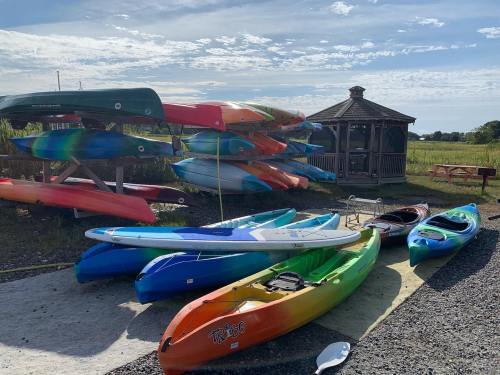 Colorful kayaks stacked on a rack, with a few on the ground, near a gazebo and grassy area under a blue sky.