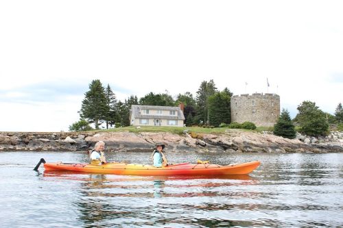 Two people kayak in calm waters near a stone castle and a house surrounded by trees.
