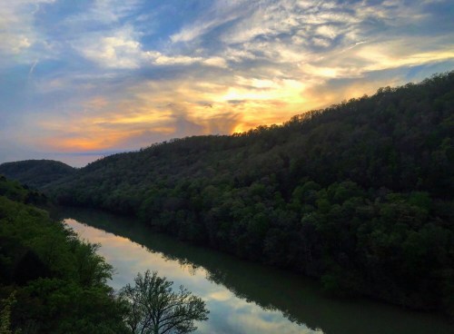 A serene river reflects a colorful sunset, surrounded by lush green hills and a cloudy sky.