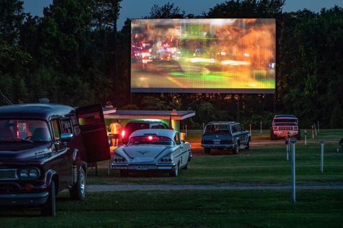 A nostalgic drive-in theater scene at dusk, featuring classic cars parked in front of a large movie screen.