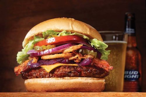 A juicy burger with lettuce, tomato, onion, pickles, and bacon, served with a beer in the background.